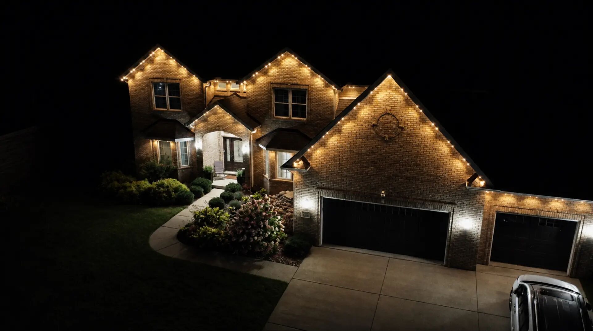 Elevated view of a brick home with warm white lighting tracing its peaks above a three-car garage